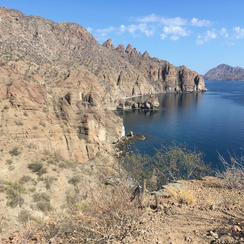 Dramatic rocky coastal cliffs dropping into a calm blue cove with a distant island on the horizon, viewed from a Loreto hiking trail