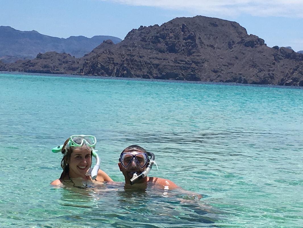 Smiling couple with snorkel masks resting in turquoise water with a dark volcanic island rising behind them in the Gulf of California near Loreto