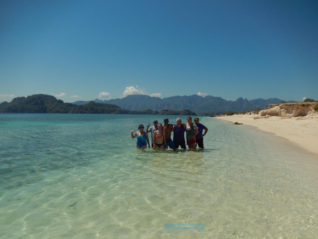 Group of six guests standing in crystal-clear water on a white sand beach with mountains behind on a Loreto island tour