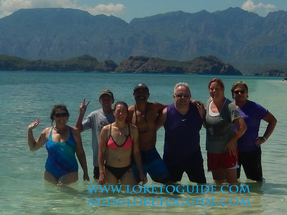 Seven happy guests standing in turquoise water with dramatic Sierra de la Giganta mountains behind on a Loreto island tour