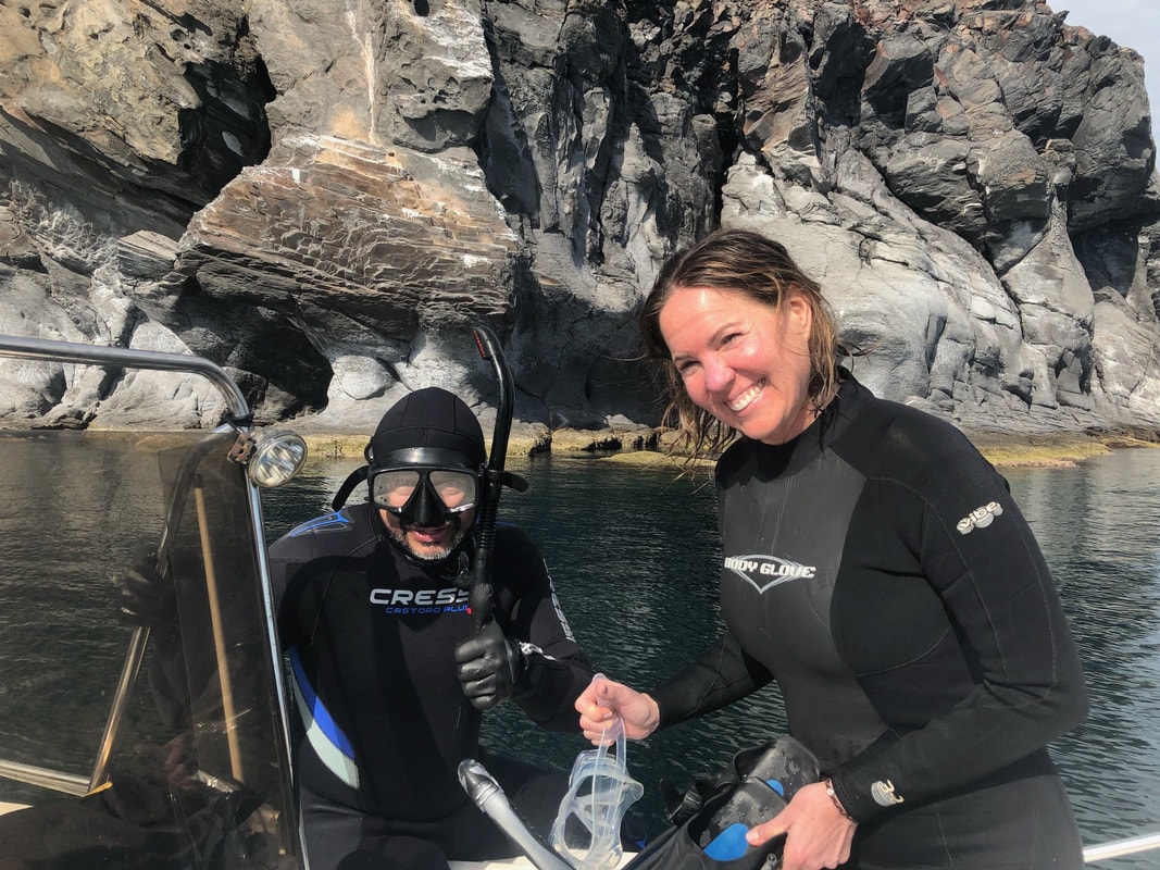 Guide in wetsuit and guest preparing snorkel gear on a boat with towering volcanic cliffs in the background near Loreto