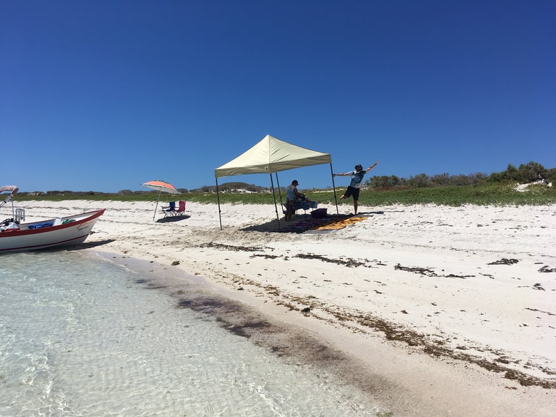 Guide jumping for joy under a shade canopy on a pristine white sand beach with a panga beached in crystal-clear water on a Loreto island tour
