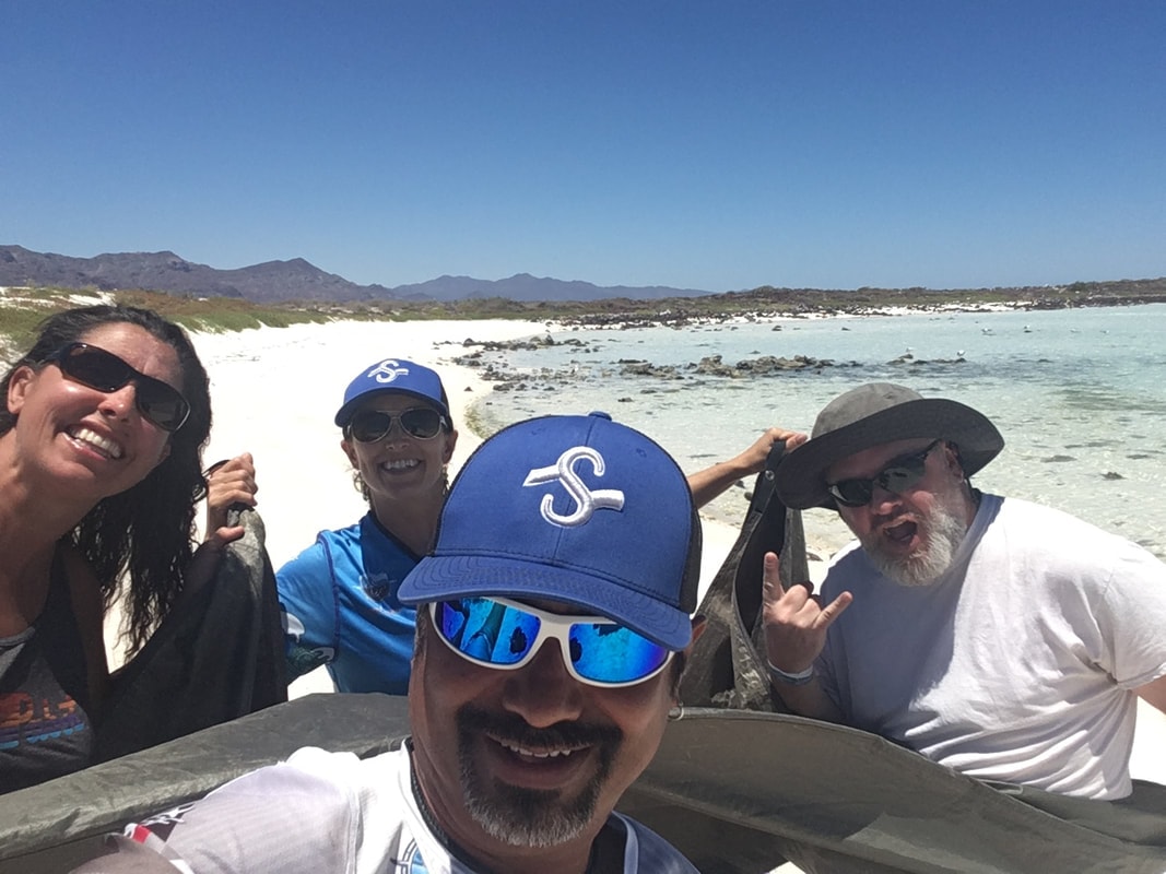 Guide taking a selfie with three happy guests on a pristine white sand beach with a turquoise lagoon and sierra mountains behind on a Loreto island tour