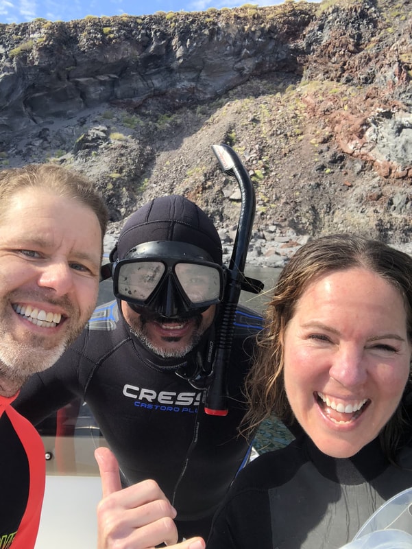 Guide in full wetsuit and mask giving a thumbs up in a selfie with two laughing guests, volcanic cliffs behind on a Loreto island tour