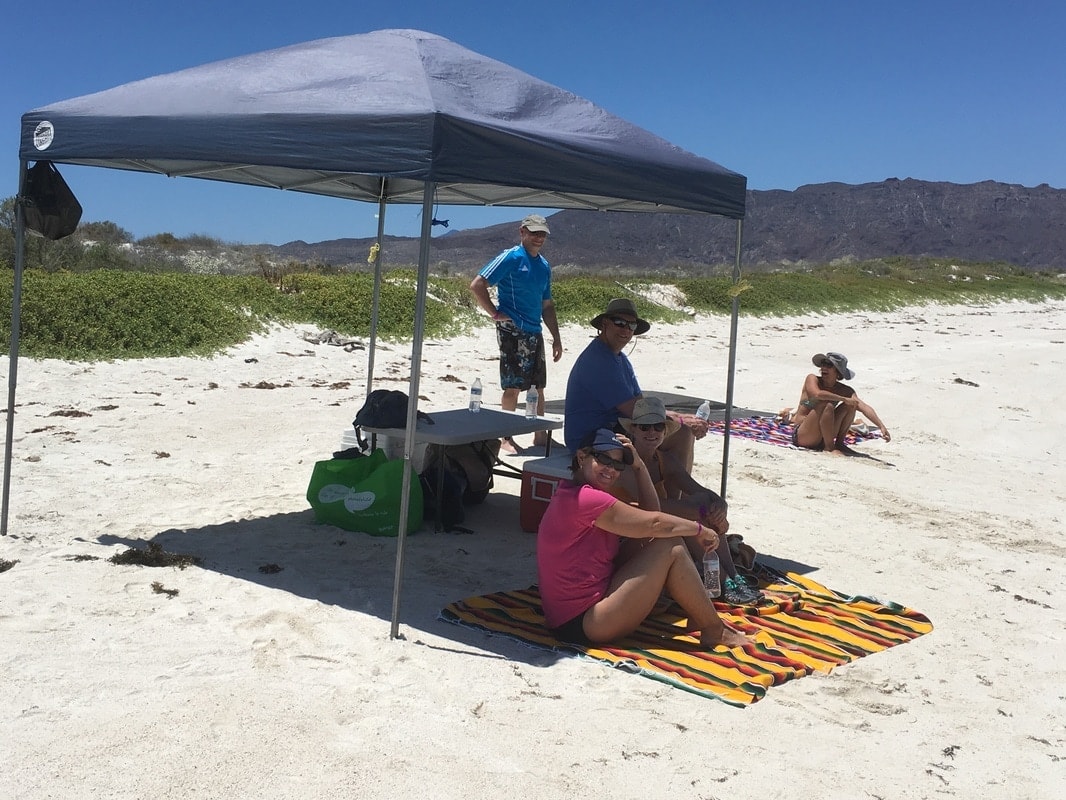 Guests relaxing under a shade canopy on a white sand beach with sierra mountains behind on a Loreto island tour