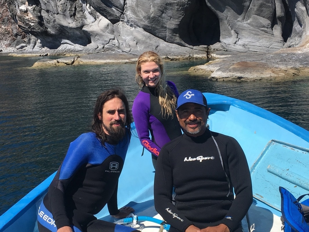 Three guests in wetsuits sitting on a blue panga with dramatic dark volcanic cave cliffs behind them on a Loreto island tour